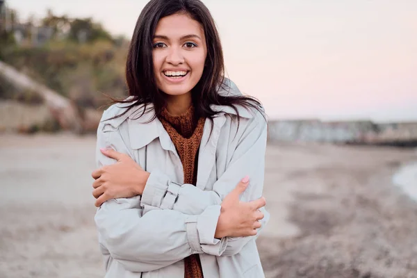 Portrett av en lystig asiatisk jente i frakk som ser glad ut i kamera mens hun går forbi havet – stockfoto