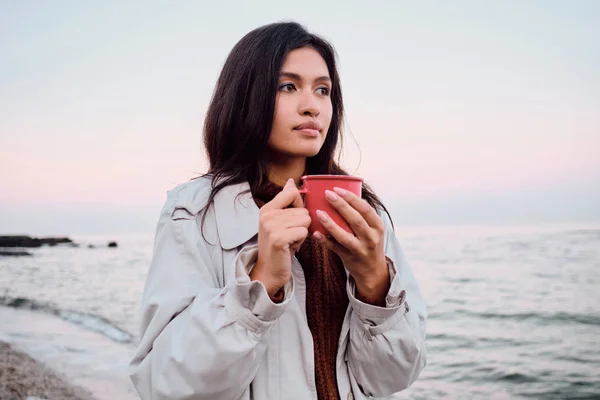 Vakker asiatisk brunette jente i frakk med en kopp varm drikk som drømmende ser bort på solnedgang strand – stockfoto