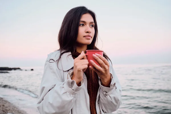 Cantik gaya gadis berambut cokelat Asia dengan jas hujan dengan secangkir minuman hangat bermimpi memandang pergi di pantai matahari terbenam — Stok Foto