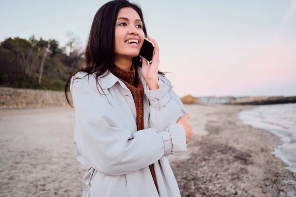 Houkutteleva hymyilevä aasialainen brunette tyttö onnellisesti puhuu matkapuhelin kävely meren rannalla — kuvapankkivalokuva