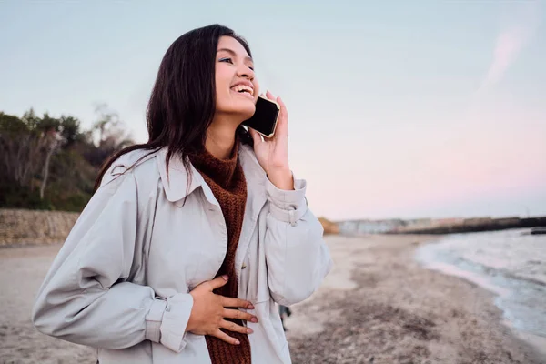 En positiv asiatisk brunette-jente som snakker i telefon på stranda. – stockfoto