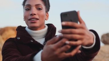 Güzel gülümseyen Afrikalı Amerikalı kız dışarıda cep telefonuyla görüntülü konuşma yaparken neşe içinde konuşuyor.