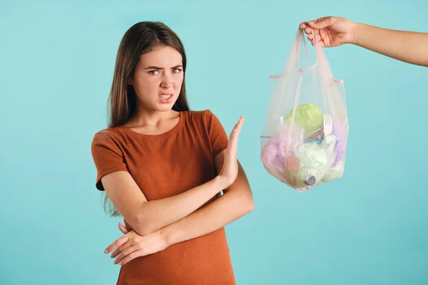 Zdenerwowana dziewczyna nie pokazuje gestu w pobliżu eko-torby z plastikowymi odpadami przyjemnie patrząc w aparacie na kolorowe tło — Zdjęcie stockowe