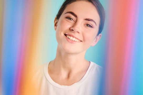 Красивая улыбающаяся девушка счастливо смотрит в камеру через радугу на красочном фоне — стоковое фото