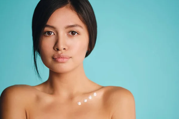 Beautiful Asian brunette girl with points of cream on clavicle intently looking in camera over colorful background — Stock Photo, Image