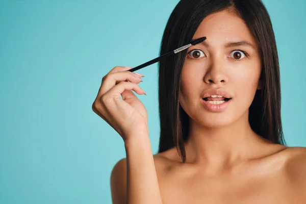 Atrakcyjne podekscytowany Azji brunetka dziewczyna z kosmetyczne szczotka stosowania żel brwi zadziwiająco patrząc w aparacie na kolorowe tło — Zdjęcie stockowe