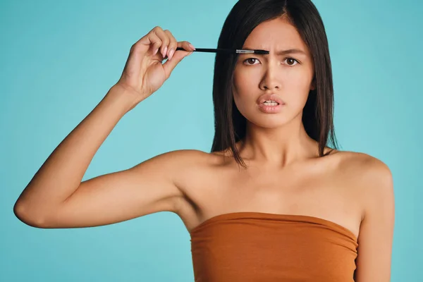 Poważny Azji brunetka dziewczyna z kosmetyczne szczotka stosowania żel brwi oburzająco patrząc w aparacie na kolorowe tło — Zdjęcie stockowe
