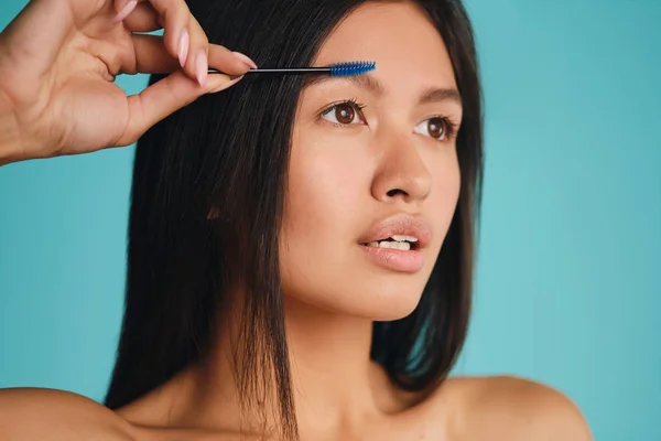 Portret atrakcyjnej azjatyckiej brunetki z pędzlem kosmetycznym starannie nakładającym żel brwiowy na kolorowe tło — Zdjęcie stockowe