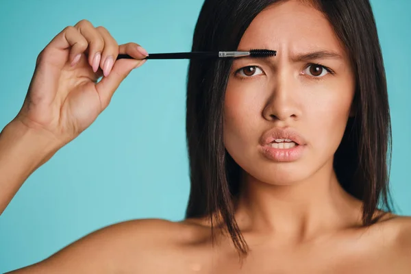 Blisko poważne Azji brunetka dziewczyna z kosmetyczne szczotka stosowania żel brwi gniewnie patrząc w aparacie na kolorowe tło — Zdjęcie stockowe
