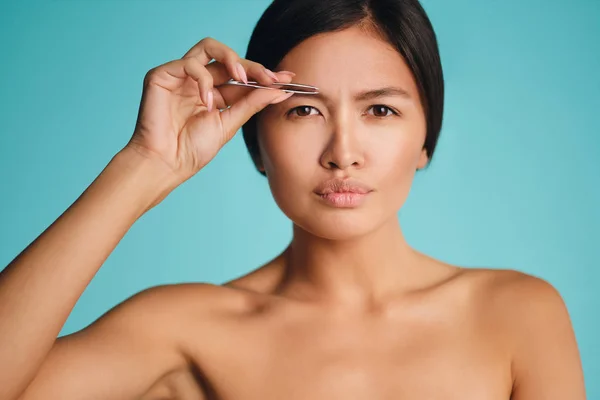 Joven hermosa mujer morena asiática concentradamente arrancando las cejas sobre fondo colorido —  Fotos de Stock