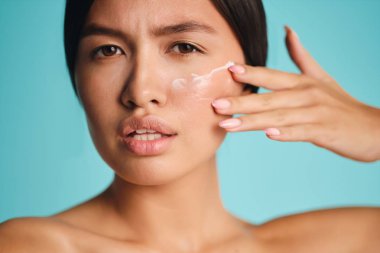 Çekici Asyalı esmer kız temiz cildine krem sürüyor. Renkli arka planda kameraya dikkatlice bakıyor.