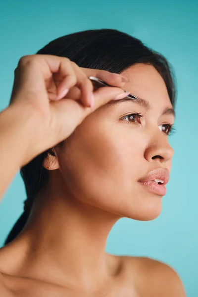 Portrait of attractive Asian brunette woman plucking eyebrows over colorful background — Stock Photo, Image