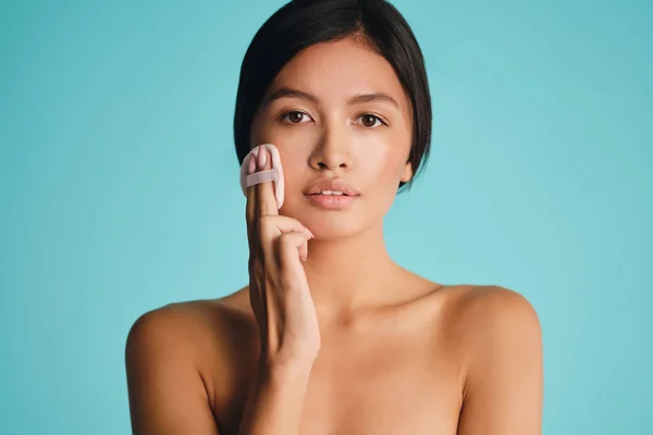 Retrato de hermosa mujer morena asiática aplicando colorete en la cara con polvo soplo sobre fondo colorido —  Fotos de Stock