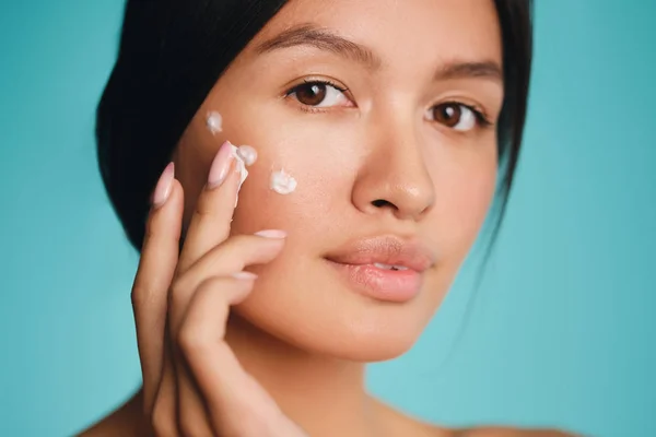 Retrato de chica morena bastante asiática con puntos de crema en la mejilla con confianza mirando en la cámara sobre fondo colorido —  Fotos de Stock