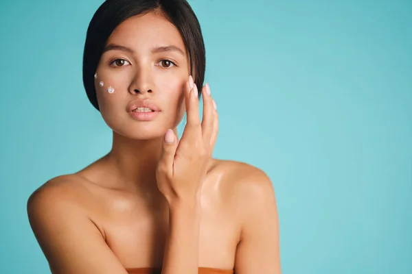 Mooi aziatisch brunette meisje met punten van crème op wang sensueel kijken in camera over kleurrijke achtergrond — Stockfoto
