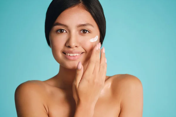 Bastante sonriente chica morena asiática aplicando crema en la piel limpia felizmente mirando en la cámara sobre fondo colorido —  Fotos de Stock