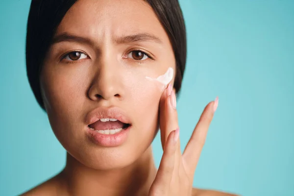 De cerca indignada chica asiática aplicando crema en la piel limpia asombradamente mirando en la cámara sobre el fondo colorido —  Fotos de Stock