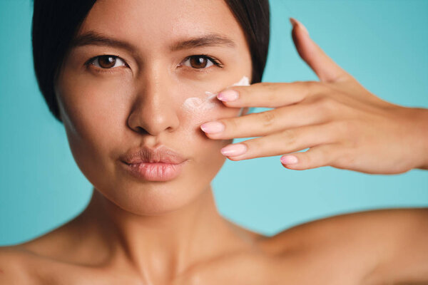 Красивая азиатская брюнетка наносит крем на чистую кожу уверенно глядя в камеру на красочном фоне
