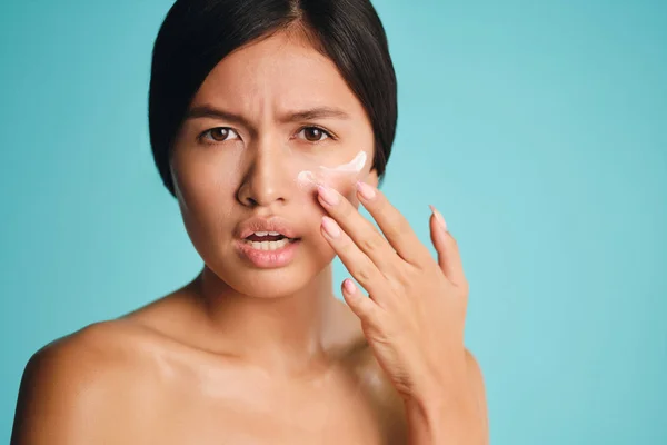 De cerca triste chica asiática aplicando crema en la piel limpia indignado mirando en la cámara sobre fondo colorido —  Fotos de Stock