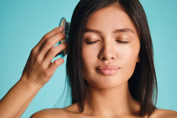 Bella ragazza bruna asiatica sognante pettinando i capelli su sfondo colorato — Foto Stock