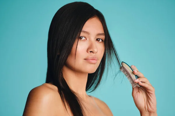 Beautiful Asian brunette girl confidently looking in camera combing hair over colorful background — Stock Photo, Image