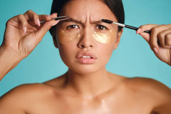 Close up Asian girl with patches under eyes seriously looking in camera with tweezers and brow brush in hands over colorful background — Stock Photo, Image