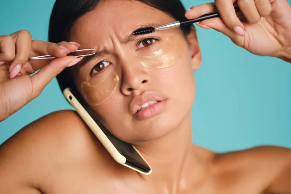 Upset Asian girl with patches under eyes talking on cellphone with tweezers and brow brush in hands over colorful background — Stock Photo, Image
