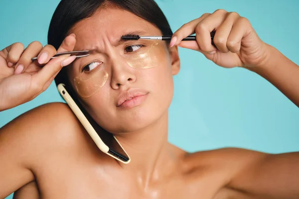 Busy Asian girl with patches under eyes putting cellphone between head and shoulder while using tweezers and brow brush over colorful background — Stock Photo, Image