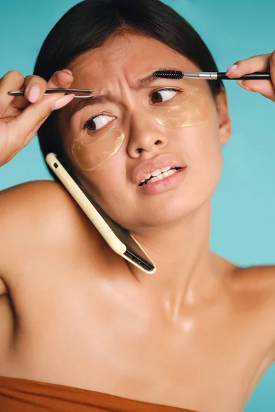 Oszołomiony Azji dziewczyna z łatami pod oczami wprowadzenie telefonu między głowę i ramię podczas korzystania z pęsety i szczotka do brwi na kolorowe tło — Zdjęcie stockowe