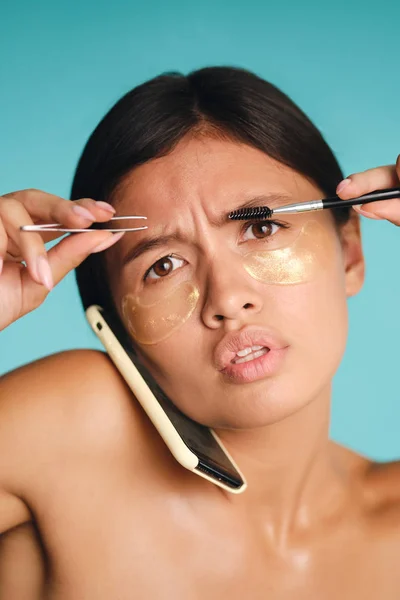 Close up Asian girl with patches under eyes talking on cellphone while using tweezers and brow brush over colorful background Royalty Free Stock Photos