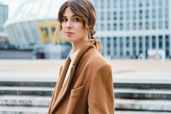 Young stylish woman in coat confidently looking in camera on city street — Stock Photo, Image