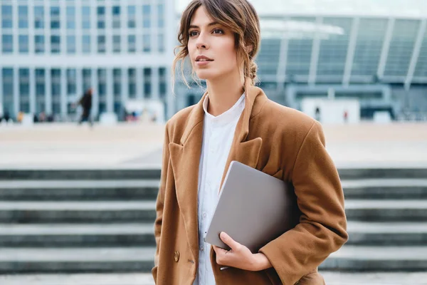 Молодая привлекательная стильная деловая женщина в пальто с ноутбуком мечтательно глядя в сторону на городской улице — стоковое фото