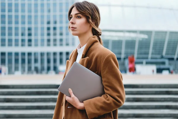 Jeune belle femme d'affaires en manteau avec ordinateur portable regardant attentivement loin sur la rue de la ville — Photo