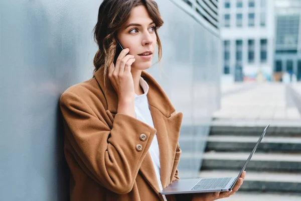 Jonge stijlvolle zakenvrouw in jas bedachtzaam praten op mobiele telefoon werken op laptop in de stad straat — Stockfoto