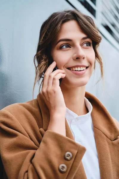 Молодая красивая стильная деловая женщина в пальто счастливо смотрит в сторону, разговаривая по мобильному телефону на открытом воздухе — стоковое фото