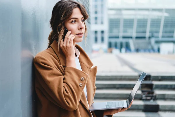 Młoda stylowa bizneswoman w płaszczu pewnie rozmawiająca przez telefon komórkowy pracująca na laptopie na ulicy miejskiej — Zdjęcie stockowe