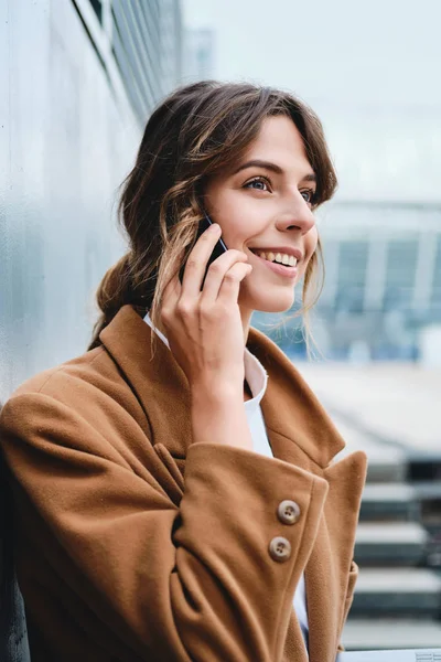 Молодая довольно стильная деловая женщина в пальто счастливо разговаривает по мобильному телефону на городской улице — стоковое фото