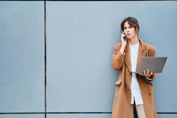 Young serious businesswoman in coat with laptop thoughtfully talking on cellphone outdoor — ストック写真