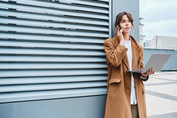 Молодая привлекательная деловая женщина в пальто уверенно разговаривает по мобильному телефону, работая на открытом воздухе — стоковое фото