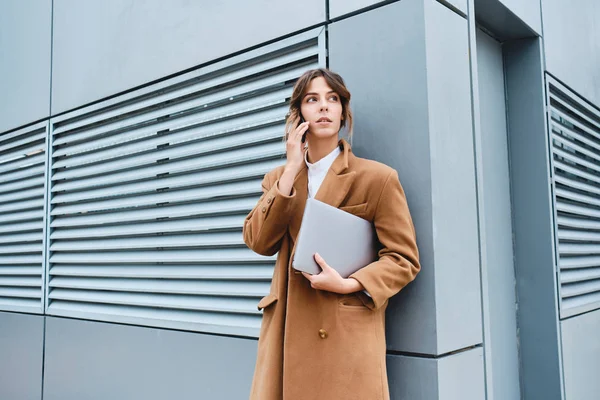 Giovane donna d'affari fiduciosa in cappotto con computer portatile parlando sul cellulare all'aperto — Foto Stock