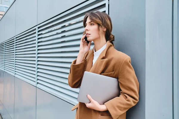 Вид сбоку на серьезную деловую женщину в пальто с ноутбуком разговаривая по мобильному телефону на открытом воздухе — стоковое фото