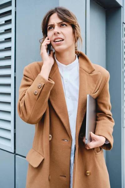 Молодая недовольная деловая женщина в пальто с ноутбуком сердито разговаривает по мобильному телефону на открытом воздухе — стоковое фото