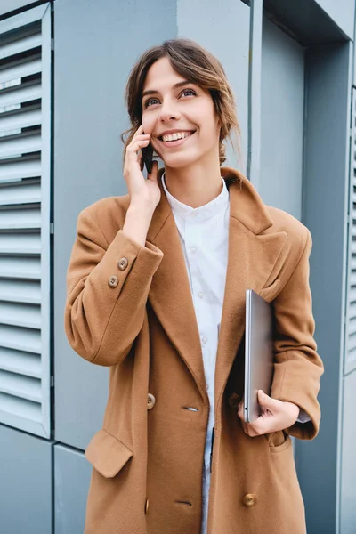 Młoda, uśmiechnięta bizneswoman w płaszczu z laptopem szczęśliwie rozmawiająca przez telefon na świeżym powietrzu — Zdjęcie stockowe