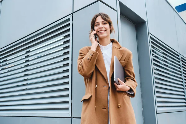Молодая красивая деловая женщина в пальто с ноутбуком радостно глядя в камеру разговаривает по мобильному телефону на открытом воздухе — стоковое фото