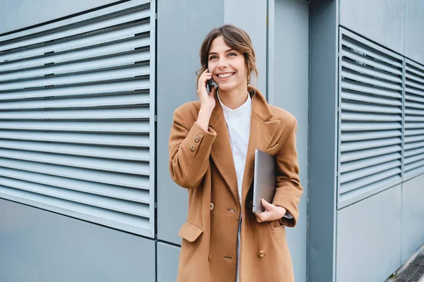 Ung, lystig forretningskvinne i frakk med bærbar pc som ser bort mens hun snakker i mobiltelefon utendørs – stockfoto
