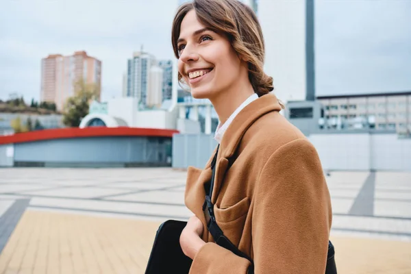 Молодая веселая стильная женщина в пальто счастливо смотрит вдаль, прогуливаясь по городской улице — стоковое фото