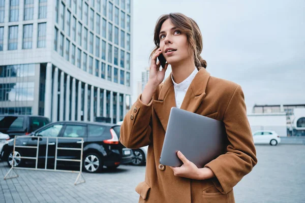 Молодая привлекательная деловая женщина в пальто с ноутбуком задумчиво разговаривает по мобильному телефону на городской улице — стоковое фото