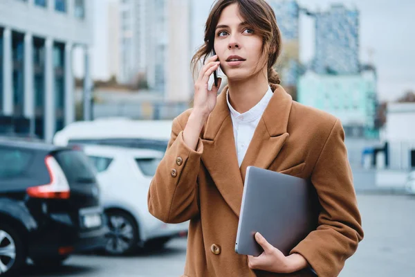 Молодая задумчивая деловая женщина в пальто с ноутбуком разговаривает по мобильному телефону пристально глядя в сторону на городской улице — стоковое фото