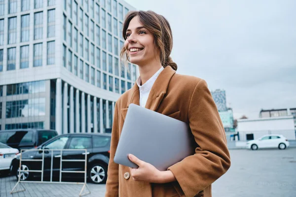 Giovane attraente donna d'affari sorridente in cappotto con computer portatile felicemente guardando lontano a piedi attraverso la strada della città — Foto Stock