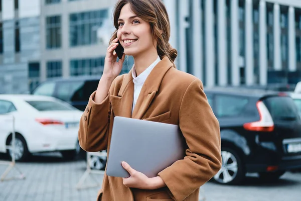 Giovane donna d'affari positiva in cappotto con computer portatile felicemente parlando sul cellulare sulla strada della città — Foto Stock